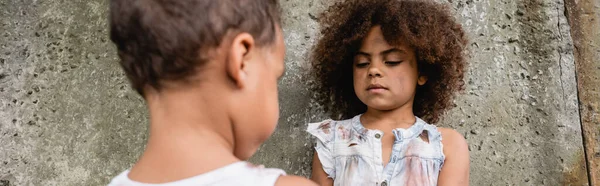 Cultivo panorámico de niño afroamericano en ropa sucia de pie cerca de hermano y muro de hormigón en la calle urbana - foto de stock