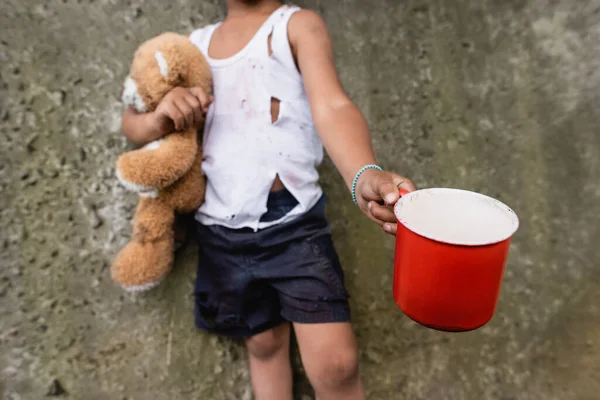 Vista recortada de chico afroamericano indigente con oso de peluche mendigando limosnas en la barriada - foto de stock