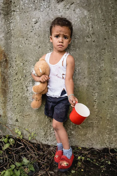 Dissatisfied african american child in dirty clothes begging alms near concrete wall in slum — Stock Photo