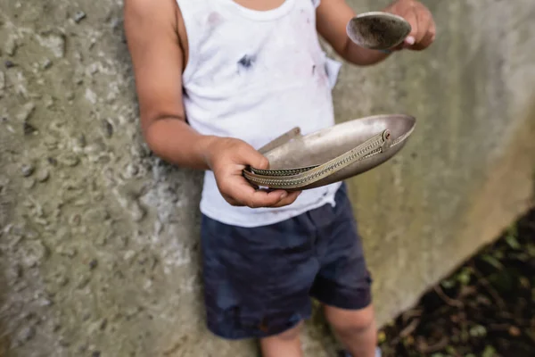 Cropped view of poor african american kid holding dirty spoon and plate on urban street — Stock Photo