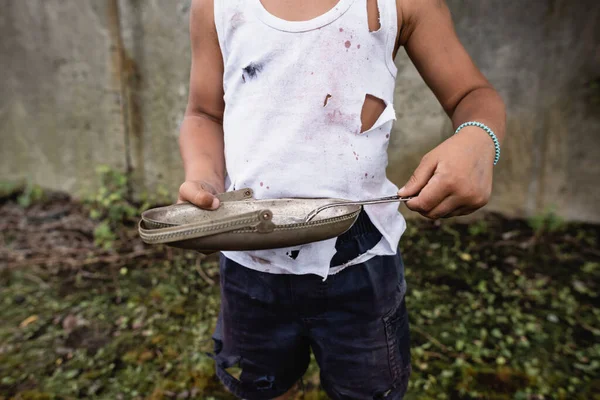 Vista recortada de un pobre niño afroamericano sosteniendo una placa de metal y una cuchara en un tugurio - foto de stock