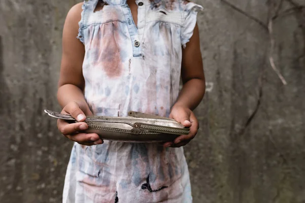 Vista recortada de un niño afroamericano pobre con ropa sucia sosteniendo placa de metal y cuchara en la calle urbana - foto de stock