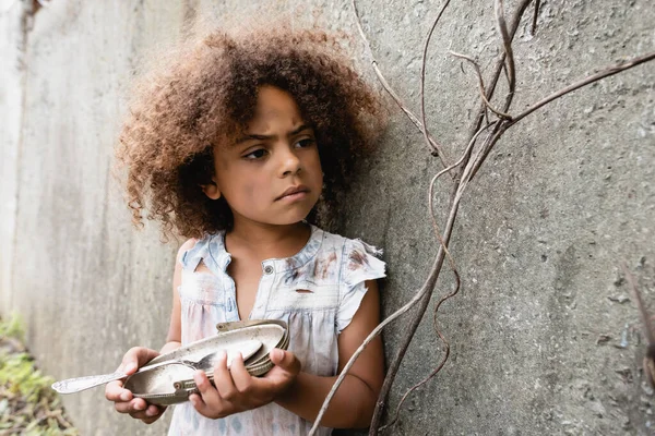 Selective focus of poor african american kid holding dirty metal plate and spoon near concrete wall on urban street — Stock Photo