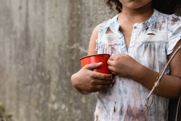 Vue recadrée d'un jeune afro-américain pauvre en vêtements sales tenant une tasse en métal dans une rue urbaine — Photo de stock