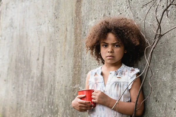 Povero bambino afroamericano che guarda la fotocamera mentre tiene in mano una tazza di metallo vicino al muro di cemento nella baraccopoli — Foto stock