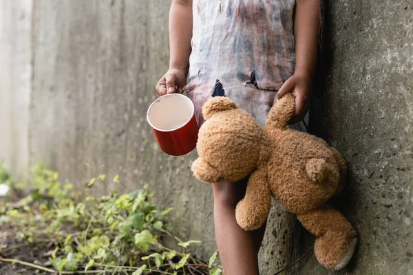 Vue recadrée d'un enfant afro-américain mendiant tenant un ours en peluche et une tasse près d'un mur de béton dans une rue urbaine — Photo de stock