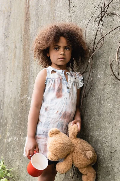 Mendigo afroamericano niño sosteniendo osito de peluche y taza cerca de muro de hormigón en la calle urbana - foto de stock