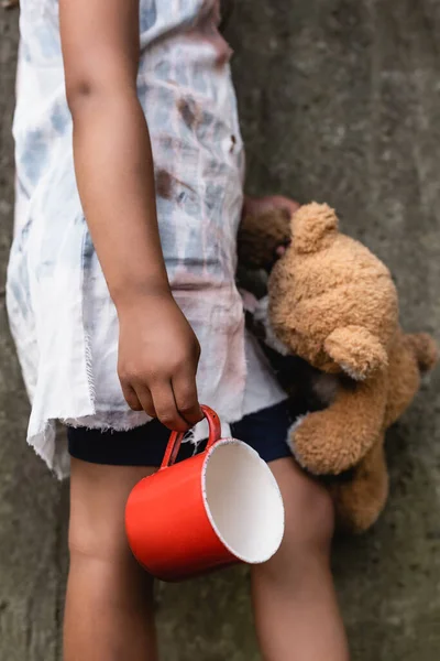 Cropped view of poor african american child holding teddy bear and metal cup on urban street — Stock Photo