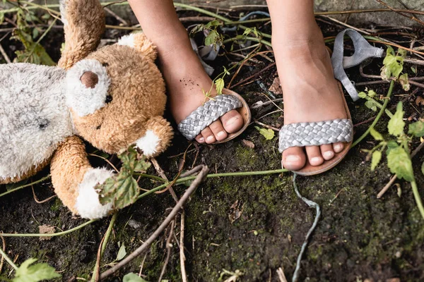 Vista recortada del oso de peluche sucio cerca de los pies de un pobre niño afroamericano en el suelo al aire libre - foto de stock
