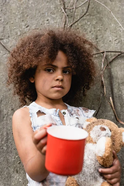 Selective focus of destitute african american child with dirty teddy bear begging alms on urban street — Stock Photo