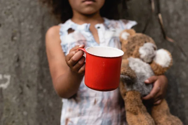 Vista ritagliata del povero bambino africano americano che tiene in mano tazza di metallo e orsacchiotto mentre implora elemosina sulla strada urbana — Foto stock