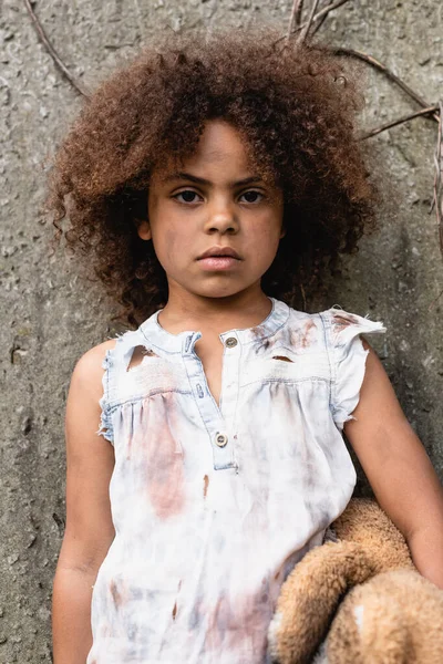 Destitute afroamericano niño sosteniendo desordenado osito de peluche y mirando a la cámara en la calle urbana - foto de stock