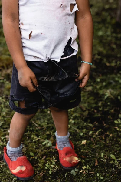 Vista recortada del pobre chico afroamericano en ropa desordenada y andrajosa de pie en la calle urbana - foto de stock