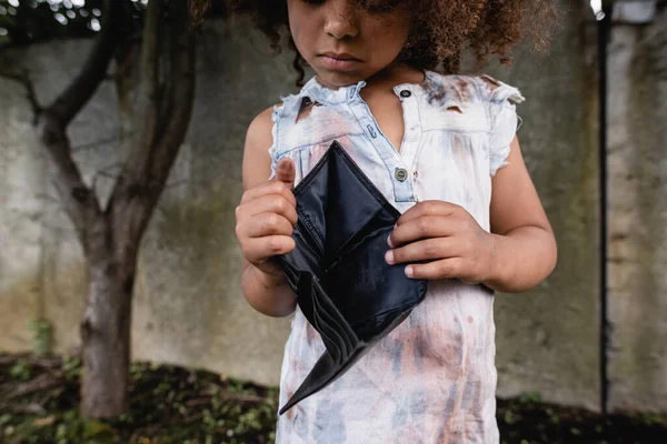 Vista recortada de un niño afroamericano pobre sosteniendo una cartera vacía en la calle urbana - foto de stock