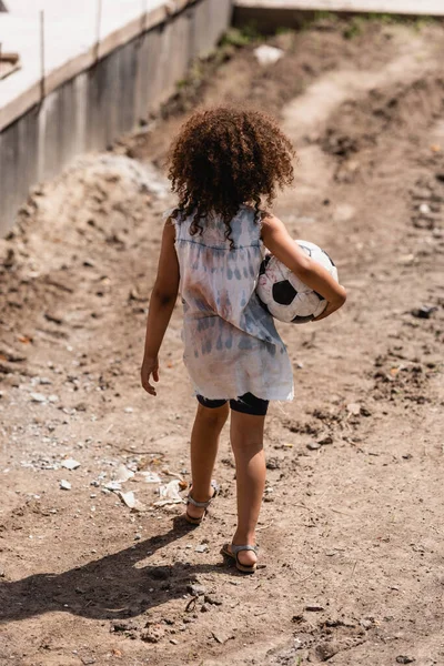 Back view of poor african american child holding soccer ball on dirty road of slum — Stock Photo