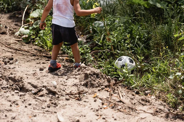 Обрізаний вид на зруйнованого афроамериканського хлопчика, що стоїть біля футбольного м'яча в траві біля брудної дороги на міській вулиці — стокове фото