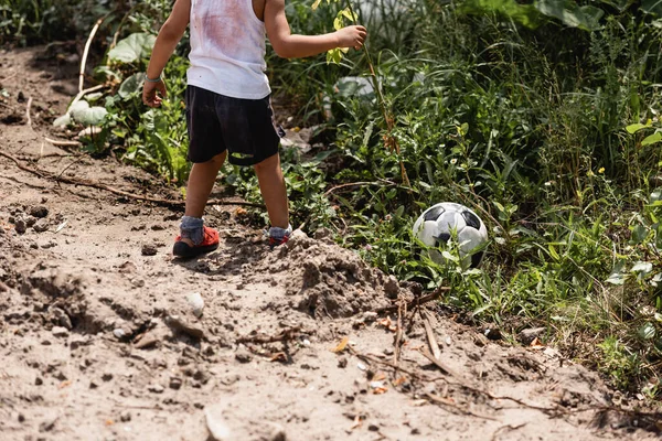 Ausgeschnittene Ansicht eines mittellosen afrikanisch-amerikanischen Jungen, der in der Nähe eines Fußballs im Gras nahe einer schmutzigen Straße auf einer städtischen Straße steht — Stockfoto