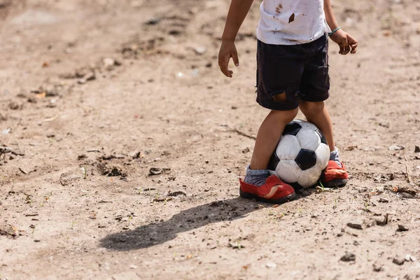 Vista recortada de un niño afroamericano indigente jugando al fútbol en el suelo en la calle urbana - foto de stock