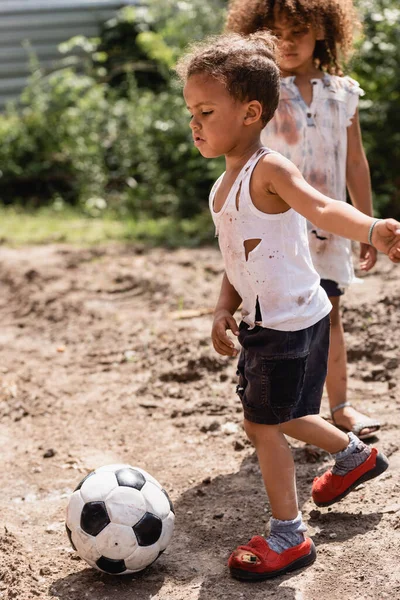 Selective focus of destitute african american boy playing football near sister on urban street in slum — Stock Photo