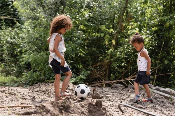 Enfoque selectivo de los niños afroamericanos indigentes que juegan al fútbol en el camino sucio en la calle urbana - foto de stock