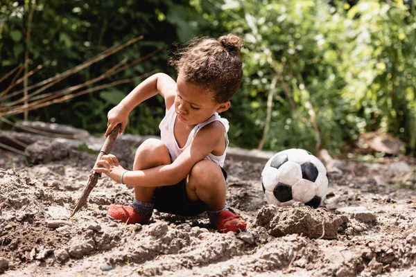 Pobre niño afroamericano sosteniendo ramita de madera cerca de pelota de fútbol en camino sucio en la calle urbana - foto de stock