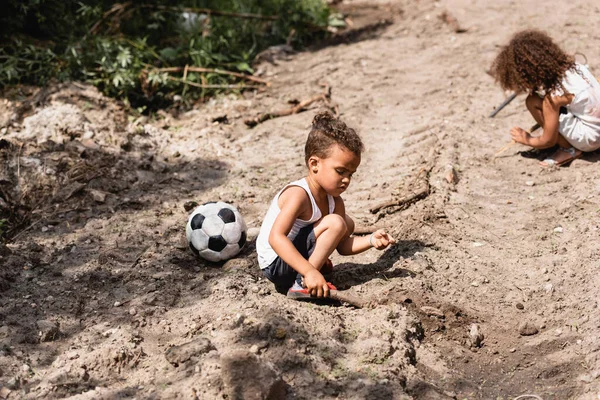 Enfoque selectivo de niños afroamericanos pobres jugando en camino sucio cerca del fútbol en la calle urbana - foto de stock