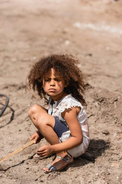Pobre niño afroamericano sosteniendo ramita de madera mientras juega en el camino sucio en la calle urbana - foto de stock