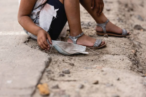 Обрезанный вид бедного африканского американского ребенка, держащего грязную медицинскую маску, сидя на земле на городской улице — стоковое фото