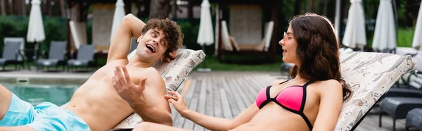 Panoramic crop of cheerful woman in sunglasses looking at handsome and shirtless man gesturing while lying on sunbed — Stock Photo