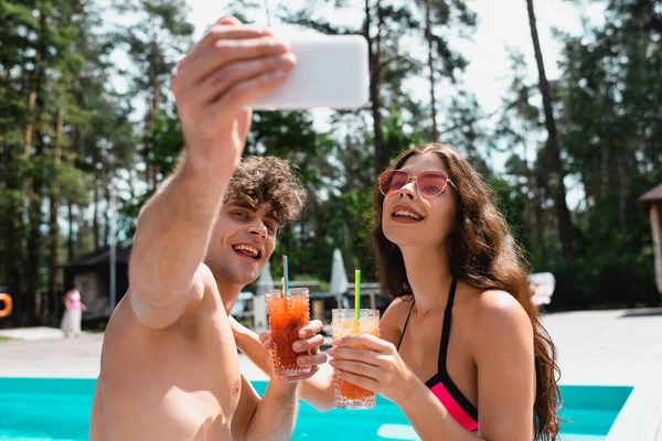 Foco seletivo de homem e mulher alegres em óculos de sol segurando coquetéis e tomando selfie — Fotografia de Stock