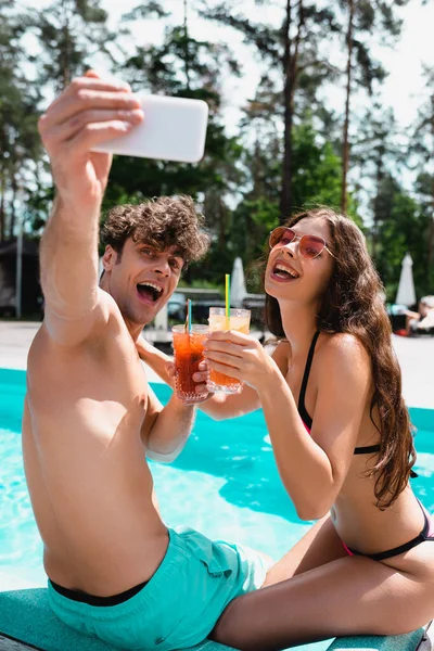 Selective focus of happy man and woman in sunglasses holding cocktails and taking selfie near swimming pool — Stock Photo