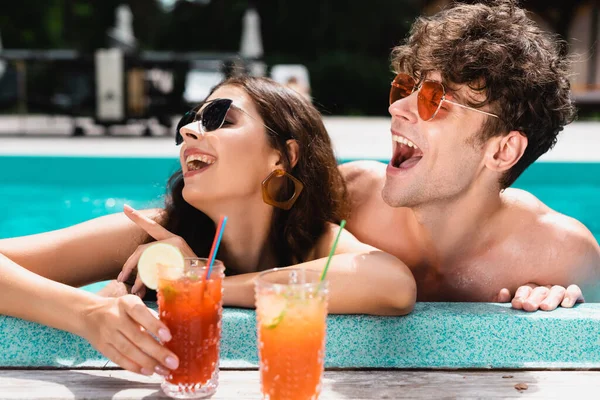 Mujer feliz en gafas de sol señalando con el dedo cerca de hombre y cócteles - foto de stock