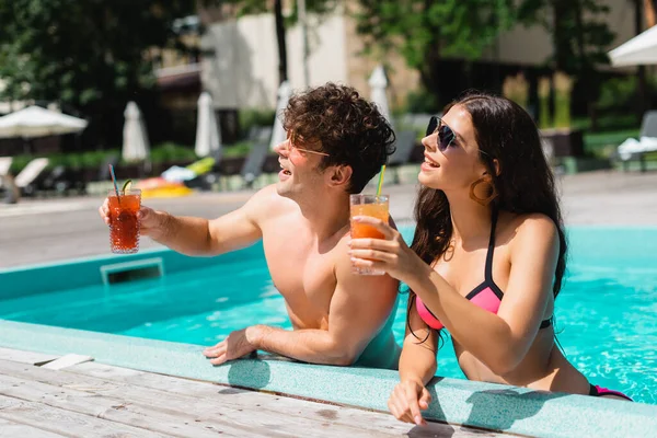 Coppia allegra in occhiali da sole che tiene cocktail in piscina — Foto stock