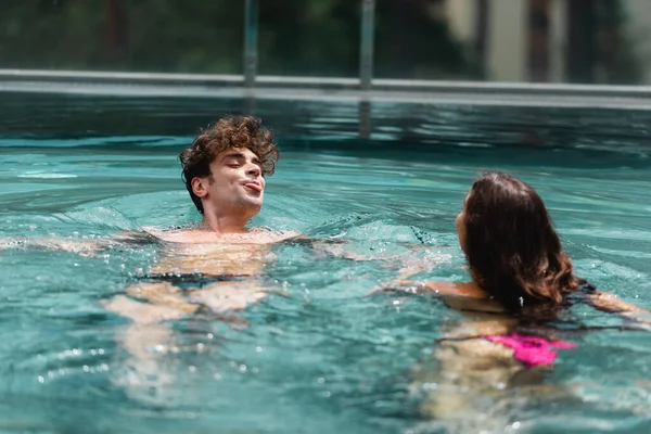 Guapo hombre sobresaliendo lengua mientras nadando con mujer en la piscina - foto de stock