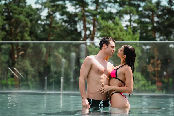Hombre musculoso abrazando y mirando a la chica en traje de baño de pie en la piscina - foto de stock