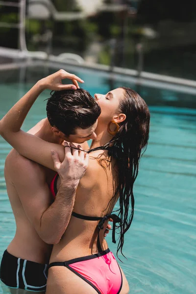 Мужчина без рубашки целует чувственную девушку в купальнике, стоящую в бассейне — стоковое фото