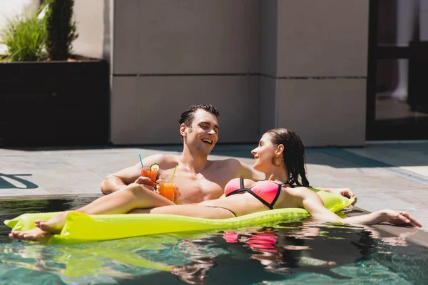 Щаслива жінка лежить на надувному матраці біля веселого чоловіка, що тримає коктейль у басейні — стокове фото