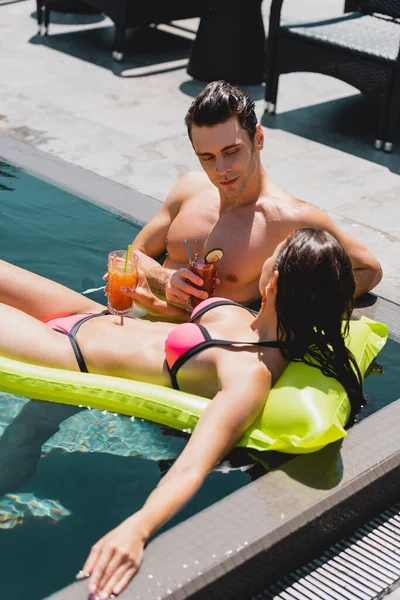 Mujer acostada en colchón inflable cerca de hombre muscular celebración de cóctel en la piscina - foto de stock