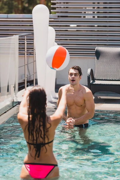 Vista trasera de la chica y el hombre sin camisa jugando voleibol con pelota de playa en la piscina - foto de stock