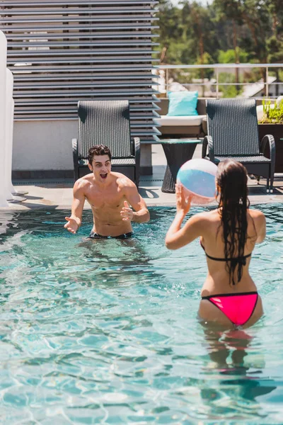 Visão traseira da mulher e homem sem camisa jogando vôlei com bola de praia na piscina — Fotografia de Stock