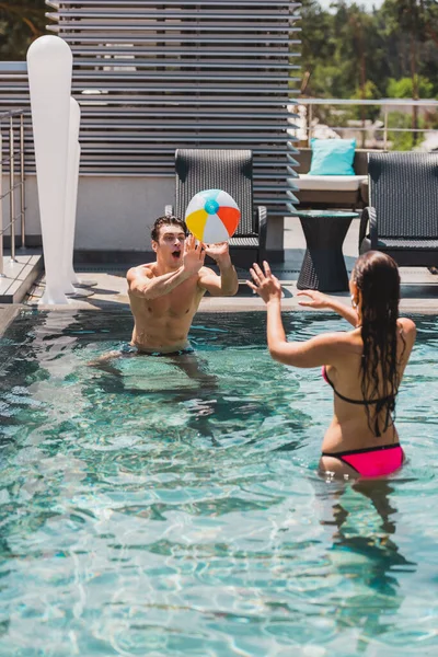 Visão traseira da mulher e homem sem camisa jogando vôlei com bola de praia na piscina — Fotografia de Stock