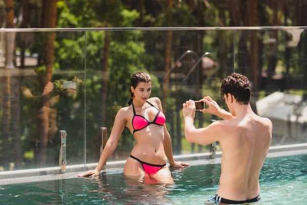 Hombre sin camisa tomando foto de chica sexy en traje de baño de pie en la piscina - foto de stock