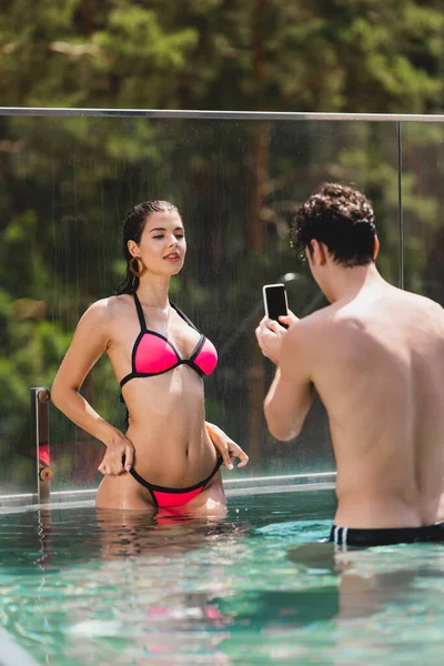 Homem muscular tirando foto de menina sexy em maiô de pé na piscina — Fotografia de Stock