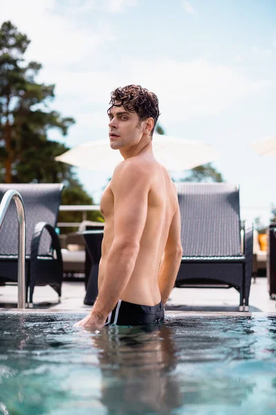 Enfoque selectivo del hombre muscular de pie en la escalera de la piscina y mirando a la cámara en la piscina - foto de stock