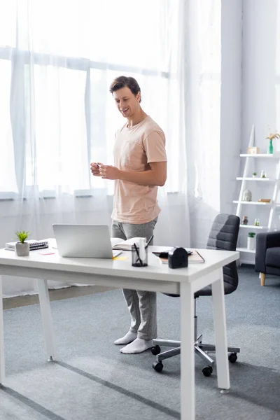 Freelancer sonriente parado frente a la computadora portátil en la mesa con papelería en casa, ganando concepto en línea - foto de stock