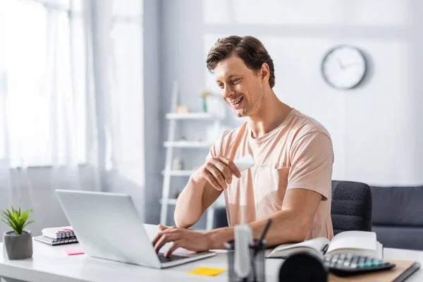 Sonriente teletrabajador mirando a la computadora portátil y sentado en la mesa con artículos de papelería en casa, ganando concepto en línea - foto de stock