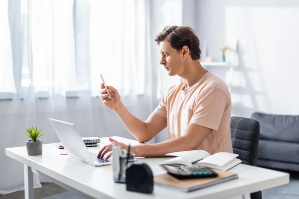 Teleworker looking at cellphone while typing on laptop, sitting on chair at home, earning online concept — Stock Photo