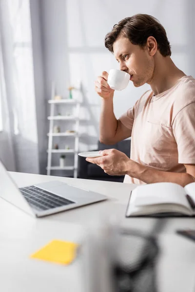 Foco seletivo de freelancer concentrado olhando para laptop enquanto bebe café em casa, ganhando conceito online — Fotografia de Stock
