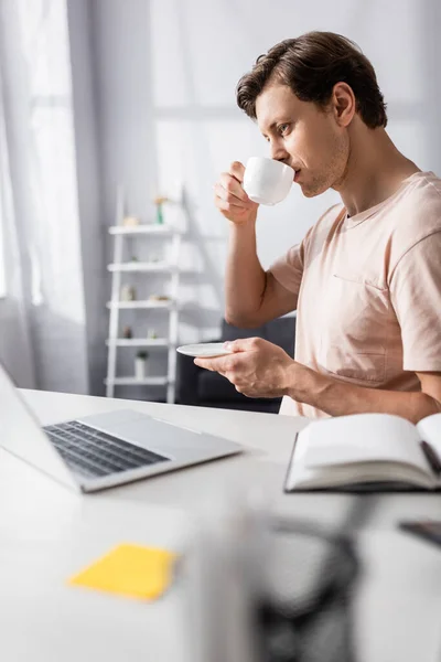 Selective focus of concentrated freelancer looking at laptop while drinking coffee at home, earning online concept — Stock Photo