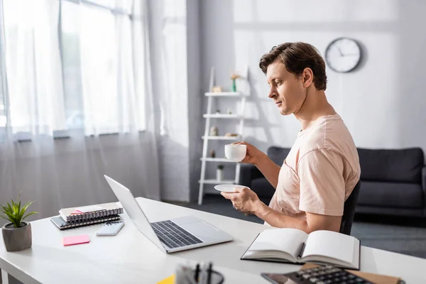 Selective focus of concentrated teleworker looking at laptop screen while drinking coffee at home, earning online concept — Stock Photo